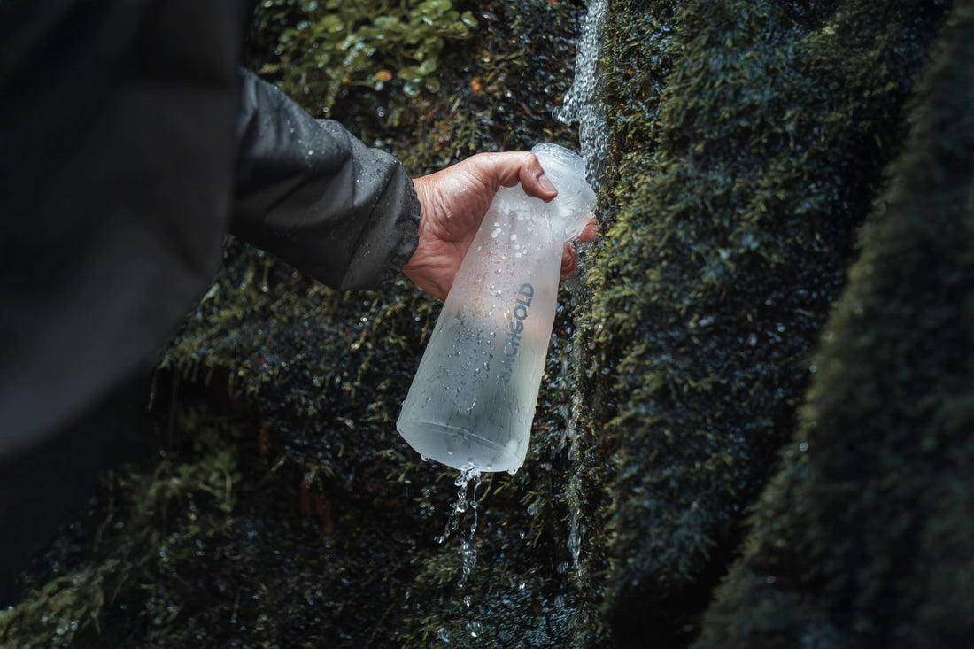 Nahaufnahme einer Hand, die eine durchsichtige Bachgold-Wasserflasche unter einen kleinen Bachlauf an moosigen Felsen hält.