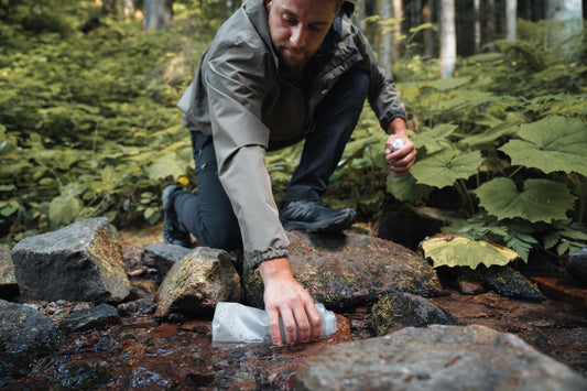 A man in hiking gear kneels on mossy rocks to fill a flexible water pouch from a shallow forest stream.