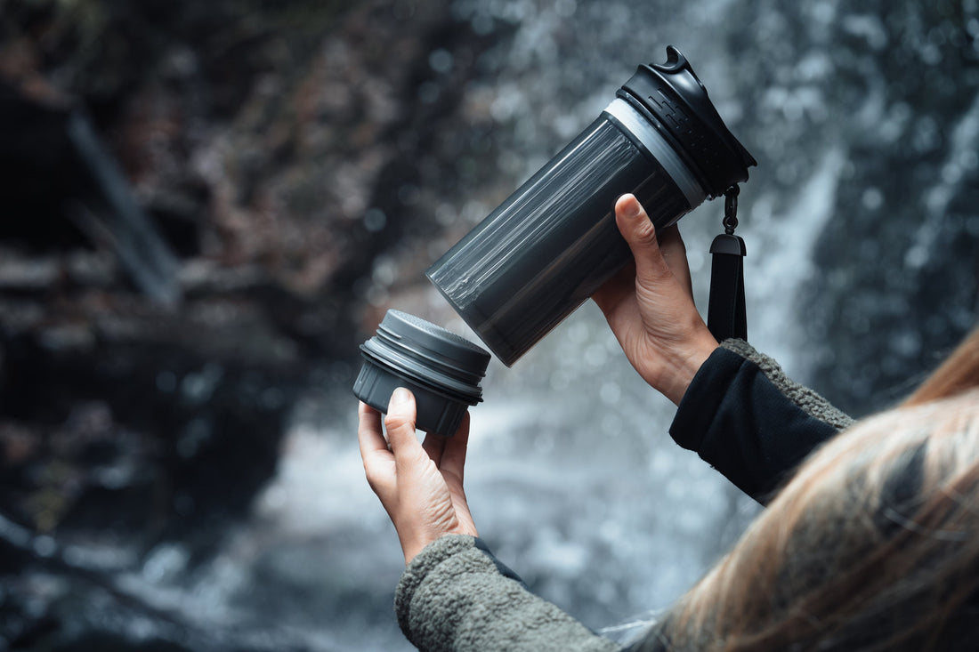 Nahaufnahme einer Person, die den Boden einer tragbaren Hohlfasermembran-Filterflasche vor einem Wasserfall abschraubt.