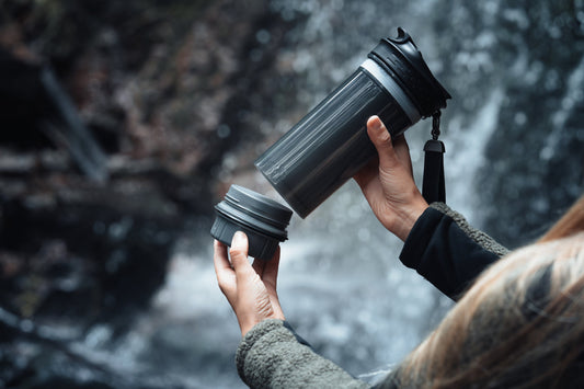 Nahaufnahme einer Person, die den Boden einer tragbaren Hohlfasermembran-Filterflasche vor einem Wasserfall abschraubt.