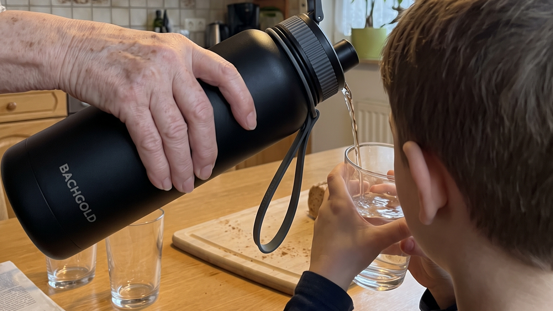 Eine Person gießt Wasser aus einer schwarzen Bachgold-Flasche in ein Glas, das ein Kind an einem Holztisch hält.