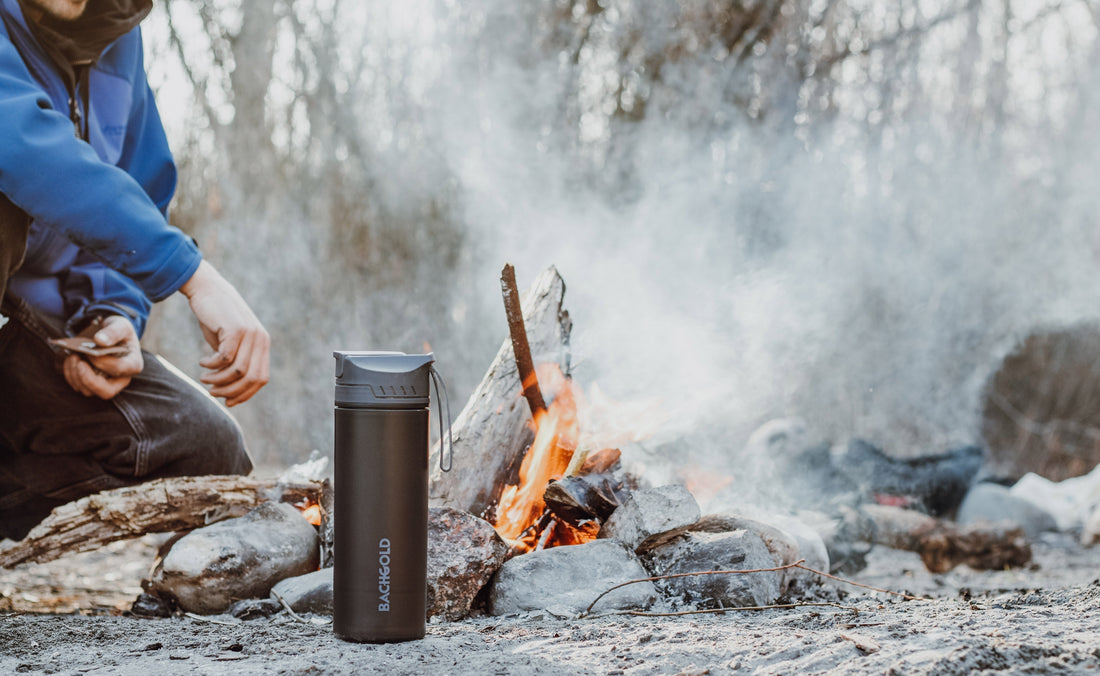 Eine schwarze Bachgold-Trinkflasche steht während einer Winterwanderung neben einem Lagerfeuer.