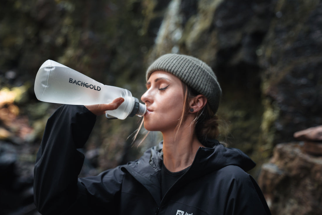 Eine Wanderin trinkt aus einer flexiblen Bachgold-Flasche vor dunkel-felsigem Hintergrund.