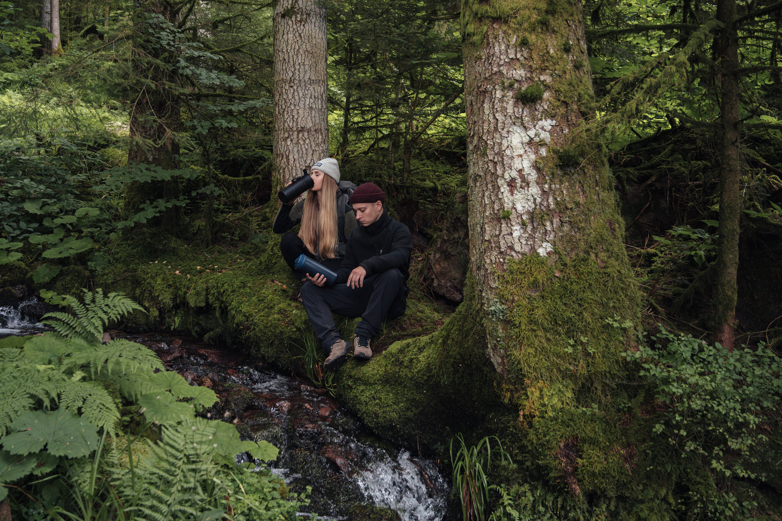 Woman drinking from Bachgold® Water Filter XL while sitting in forest with man